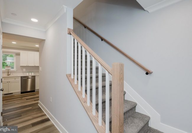 Staircase with light wood handrail and white balusters beside a hallway leading to a white kitchen with stainless appliances.