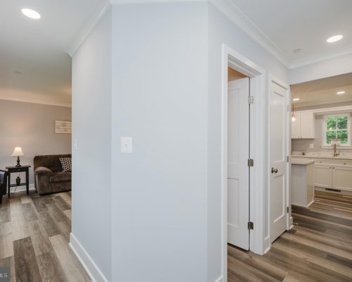Bright hallway with light gray walls and wood-look flooring, opening to a living room on the left and a white kitchen on the right.