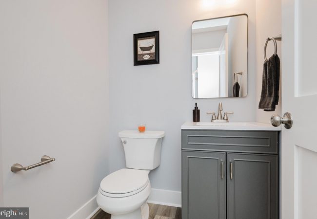 Small bathroom with a gray cabinet vanity, white sink, mirror, and toilet against pale walls.