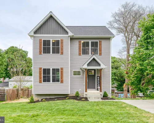 Two-story gray house with wooden shutters, a small front porch, and a vertical 'WELCOME' sign beside the door.
