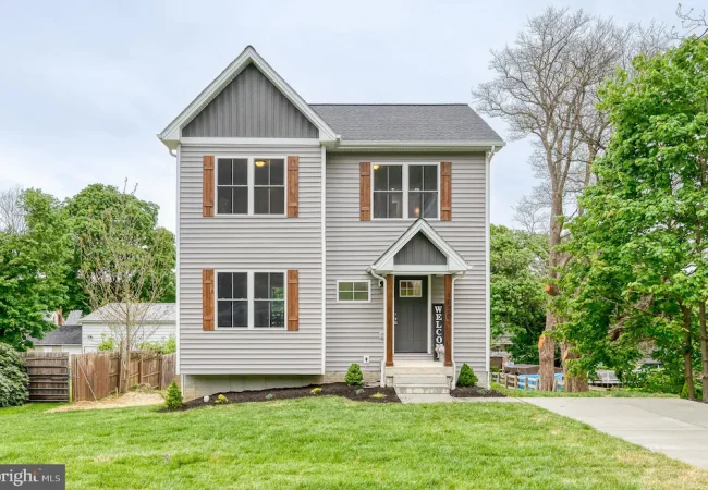 Two-story gray house with wooden shutters, a small front porch, and a vertical 'WELCOME' sign beside the door.