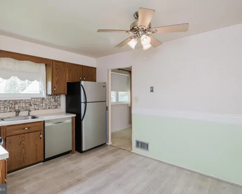Kitchen with wooden cabinets, stainless steel fridge and dishwasher, double sink under a window, and a patterned backsplash behind the counters under a ceiling fan.