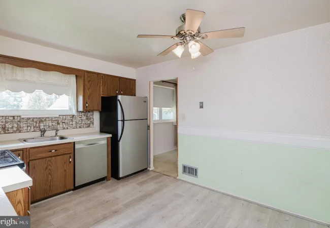 Kitchen with wooden cabinets, stainless steel fridge and dishwasher, double sink under a window, and a patterned backsplash behind the counters under a ceiling fan.