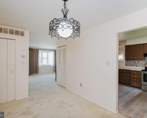 Living area with a decorative metal chandelier, cream carpet, and a doorway leading to a kitchen with wooden cabinets and stainless appliance.