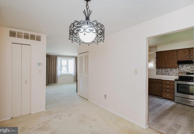 Living area with a decorative metal chandelier, cream carpet, and a doorway leading to a kitchen with wooden cabinets and stainless appliance.