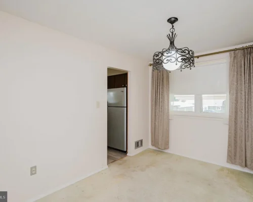 Empty living/dining room with beige walls, ornate black chandelier, and a window with taupe curtains; open doorway reveals a stainless steel refrigerator in the adjacent kitchen.