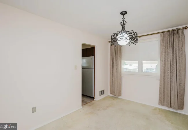 Empty living/dining room with beige walls, ornate black chandelier, and a window with taupe curtains; open doorway reveals a stainless steel refrigerator in the adjacent kitchen.