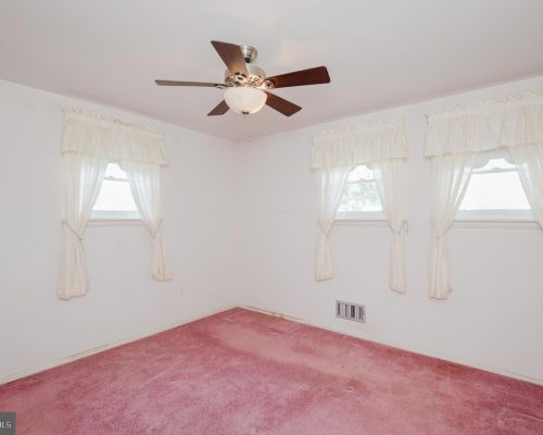 Small empty bedroom with pink carpet, white walls, and three windows with lace curtains and a ceiling fan with lights.
