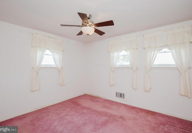 Small empty bedroom with pink carpet, white walls, and three windows with lace curtains and a ceiling fan with lights.