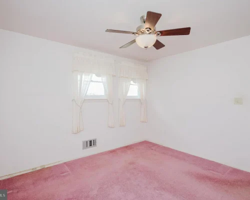 Empty bedroom with white walls and pink carpet, a ceiling fan, two small windows with lace curtains, and a doorway to an adjoining room, plus a wall vent near the baseboard.