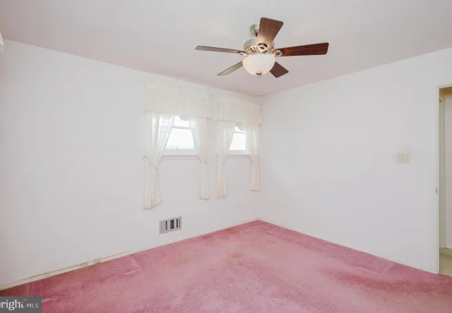 Empty bedroom with white walls and pink carpet, a ceiling fan, two small windows with lace curtains, and a doorway to an adjoining room, plus a wall vent near the baseboard.