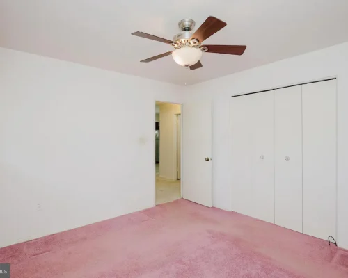 Empty bedroom with white walls, pink carpet, and a ceiling fan; large white bi-fold closet doors on one wall.