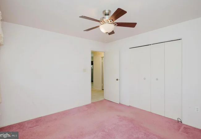 Empty bedroom with white walls, pink carpet, and a ceiling fan; large white bi-fold closet doors on one wall.