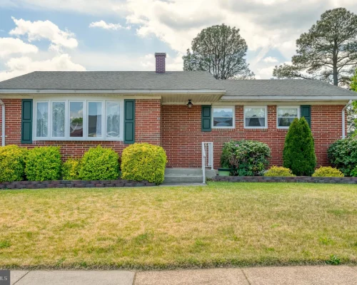 Brick single-story house with green shutters, a gray roof, and a row of trimmed boxwoods in the front yard.
