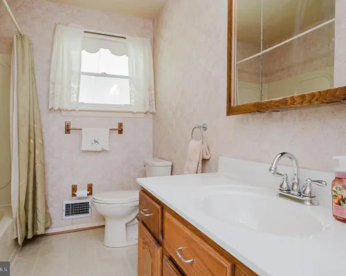 Bright pink bathroom with a white countertop sink, wooden vanity, chrome faucet, toilet, shower with a light curtain, and a window with lace curtains.