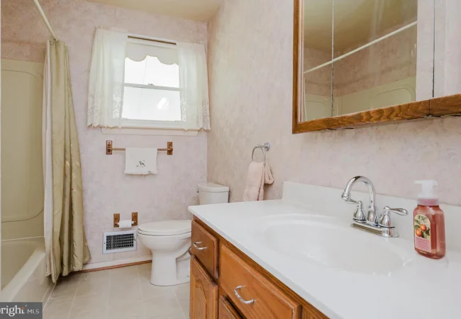 Bright pink bathroom with a white countertop sink, wooden vanity, chrome faucet, toilet, shower with a light curtain, and a window with lace curtains.
