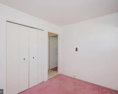 Plain white bedroom with pink carpet, a two-door closet on the left and an open doorway to a hallway on the far wall, plus a window with blue valances on the right corner.