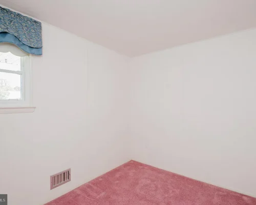 Empty corner of a small room with pink carpet, white walls, and a window with a blue valance.