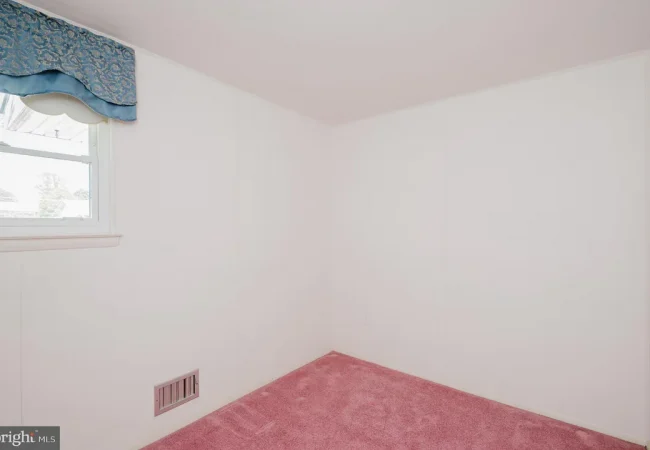 Empty corner of a small room with pink carpet, white walls, and a window with a blue valance.