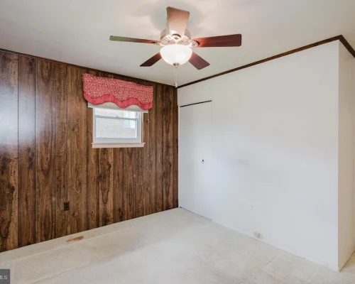 Room with a wood-paneled accent wall, small window with pink valance, ceiling fan, and white walls; beige carpet floor, closet door on the back wall.