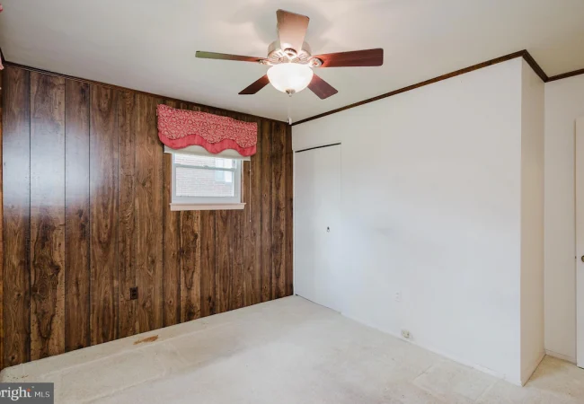 Room with a wood-paneled accent wall, small window with pink valance, ceiling fan, and white walls; beige carpet floor, closet door on the back wall.