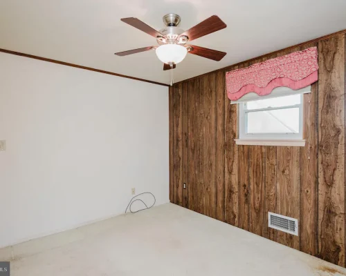 Empty room with white walls and a wood-paneled accent wall, featuring a ceiling fan with lights and a small window with a pink valance.