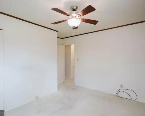 Empty bedroom with white walls, dark wood trim, and a ceiling fan; doorway to hall and closet on the left with a loose cable on the carpet outside the outlet.