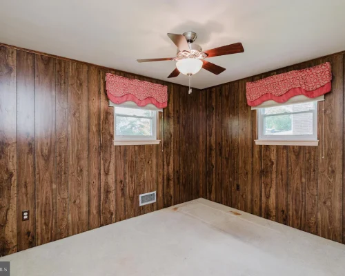 Basement-style room with dark wood paneling, two small windows with pink valances, a ceiling fan, and a stained beige carpet.