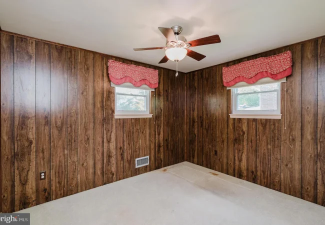 Basement-style room with dark wood paneling, two small windows with pink valances, a ceiling fan, and a stained beige carpet.