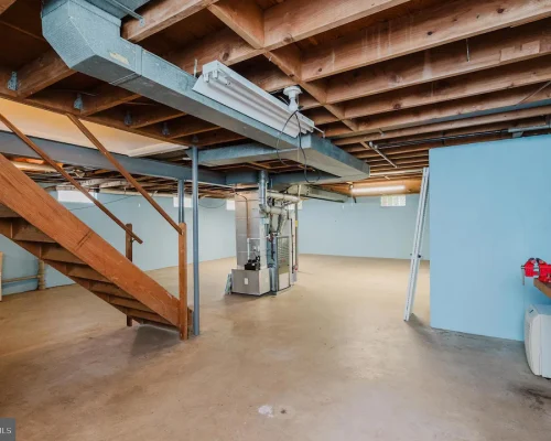 Unfinished basement with wooden stairs, exposed beams, and utility equipment along a blue wall, concrete floor, and a workbench on the right side.