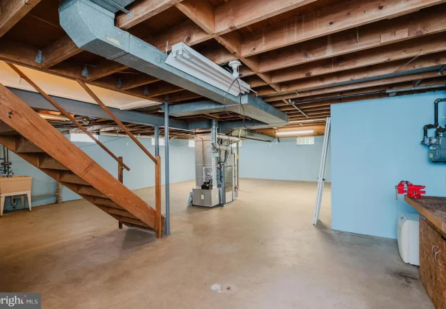 Unfinished basement with wooden stairs, exposed beams, and utility equipment along a blue wall, concrete floor, and a workbench on the right side.