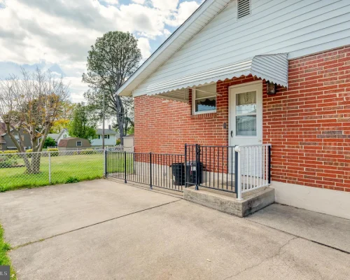 Brick house with small front porch, white door, and black railing; concrete driveway and chain-link fence in a suburban yard.