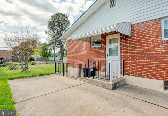 Brick house with small front porch, white door, and black railing; concrete driveway and chain-link fence in a suburban yard.