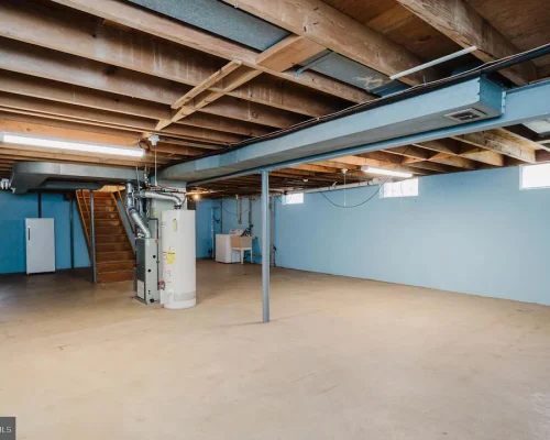 Unfinished basement with blue walls, exposed ceiling joists, a water heater and furnace, and a wooden staircase.
