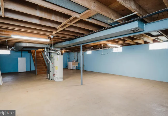 Unfinished basement with blue walls, exposed ceiling joists, a water heater and furnace, and a wooden staircase.