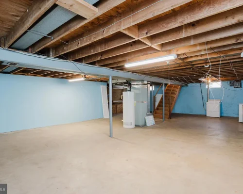 Unfinished basement with blue walls, exposed wooden ceiling beams, and a water heater near the back with laundry appliances along the far wall.