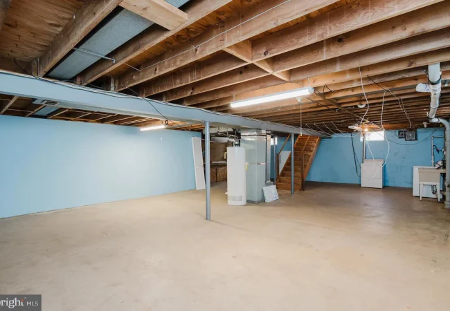 Unfinished basement with blue walls, exposed wooden ceiling beams, and a water heater near the back with laundry appliances along the far wall.