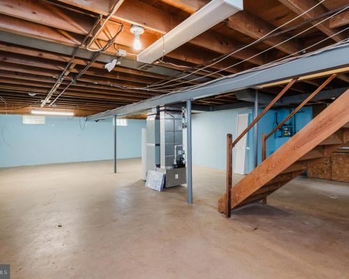 Basement with concrete floor, blue walls, exposed ceiling, and a wooden staircase on the right side leading upstairs.