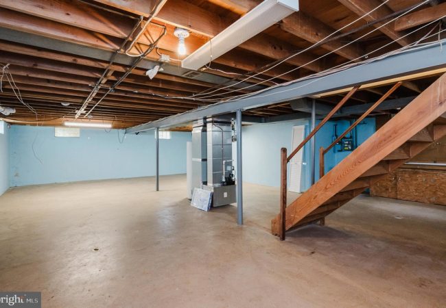 Basement with concrete floor, blue walls, exposed ceiling, and a wooden staircase on the right side leading upstairs.