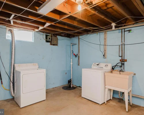 Basement laundry area with a washer, a dryer, and a utility sink against blue walls and exposed ceiling beams.