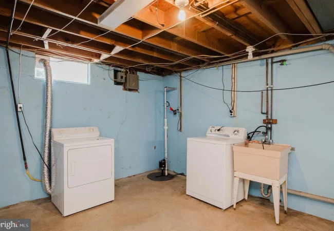 Basement laundry area with a washer, a dryer, and a utility sink against blue walls and exposed ceiling beams.