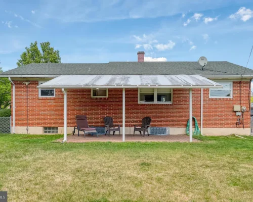 Brick house rear with a covered patio; four lawn chairs and a small table on a brick floor, green lawn beyond.