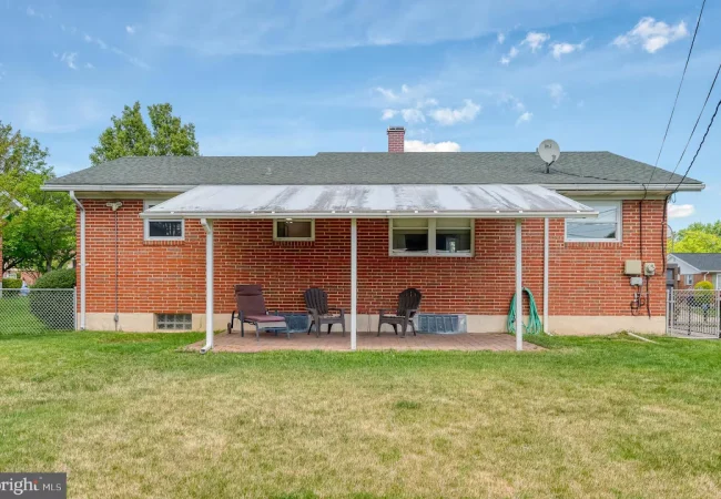 Brick house rear with a covered patio; four lawn chairs and a small table on a brick floor, green lawn beyond.
