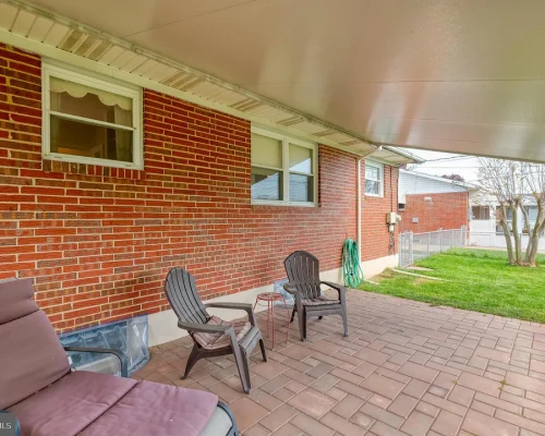Covered brick-backyard patio with brown cushioned lounge chair and two plastic chairs on a paved surface; a green hose coiled on the wall and a grassy yard beyond a chain-link fence.