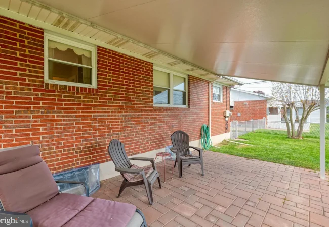 Covered brick-backyard patio with brown cushioned lounge chair and two plastic chairs on a paved surface; a green hose coiled on the wall and a grassy yard beyond a chain-link fence.