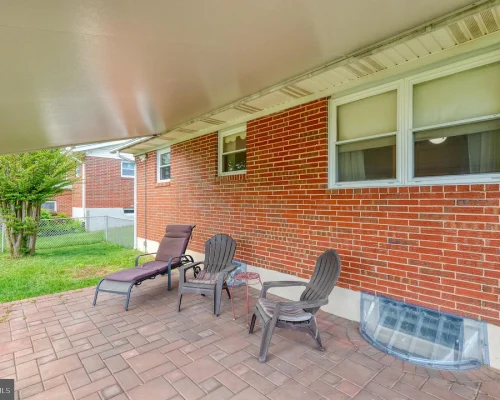 Covered brick patio with four outdoor chairs and a lounge chair along a red brick house, facing a fenced yard.