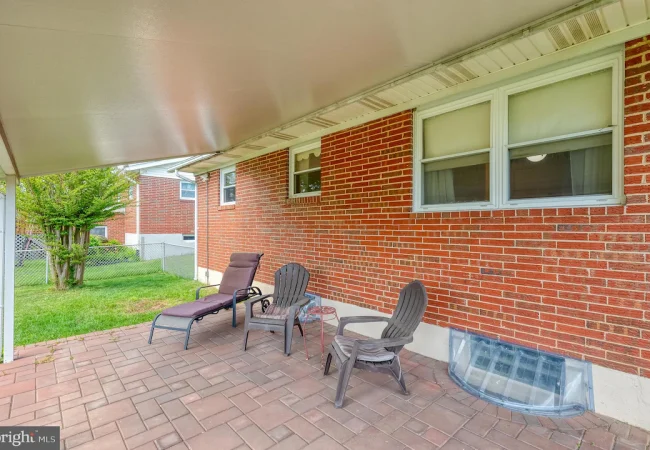 Covered brick patio with four outdoor chairs and a lounge chair along a red brick house, facing a fenced yard.