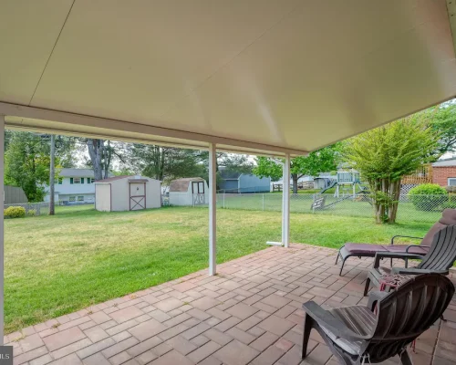 Covered patio overlooking a green backyard with lawn, sheds, and a chain-link fence; outdoor chairs and a lounge chair on brick patio.