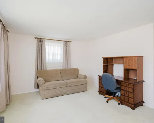 A small living room with a beige sofa, light walls, and a wooden desk with a blue rolling chair against the right wall, under a window with gray curtains.