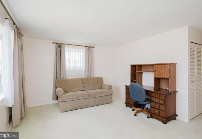 A small living room with a beige sofa, light walls, and a wooden desk with a blue rolling chair against the right wall, under a window with gray curtains.